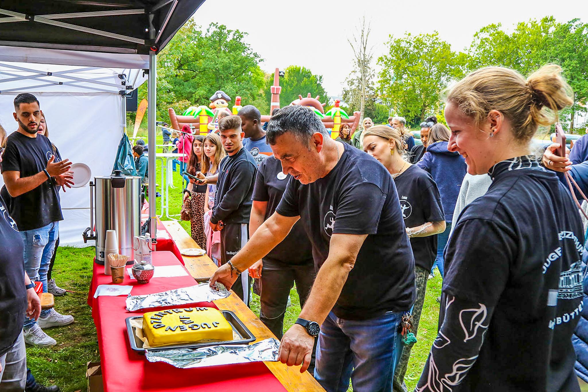 Mann schneidet gelbe Jubiläumstorte mit Aufschrift am Stand an, Menschen in dunklen T-Shirts und Besucher stehen daneben im Freien.