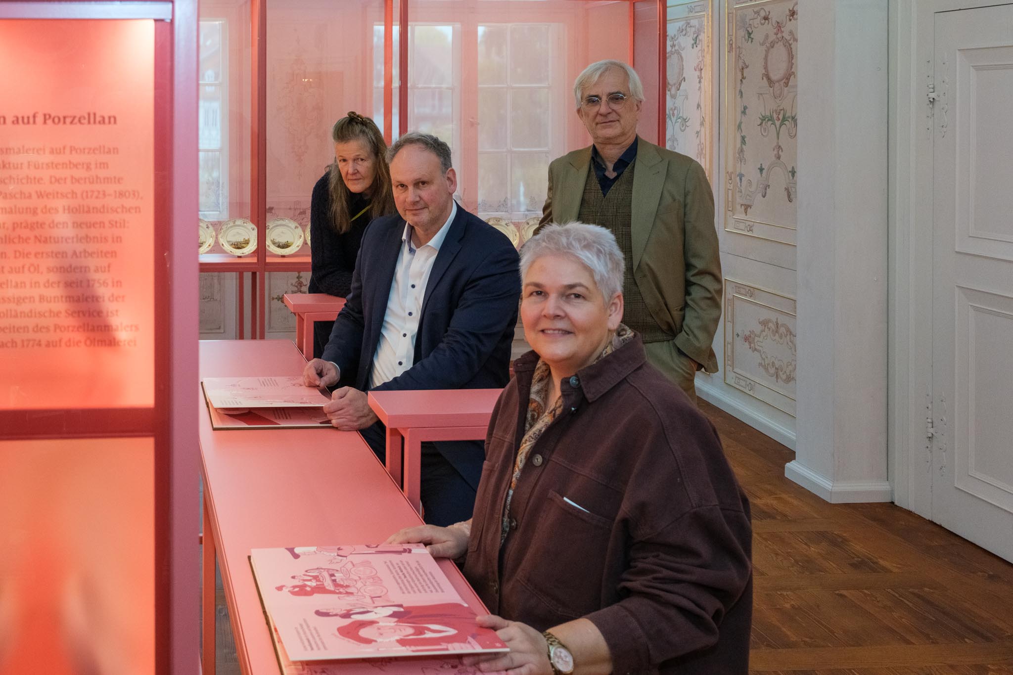 Vier Personen stehen und sitzen an pinken Ausstellungstischen im neu gestalteten Porzellankabinett, im Hintergrund Vitrinen mit Porzellantellern.