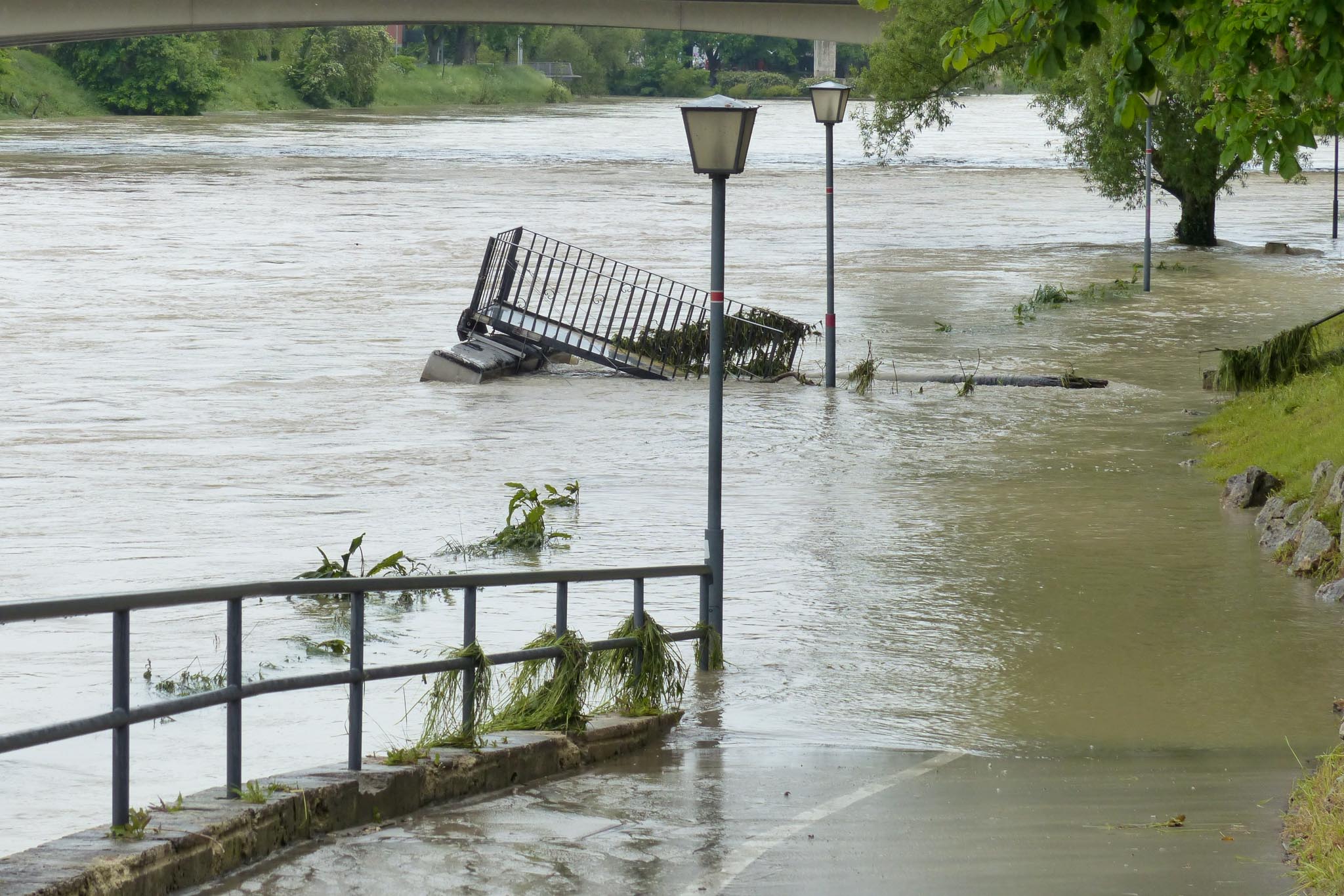 Hochwasser an einem Fluss überflutet Ufer und Wege, eine umgestürzte Treppe mit Geländer treibt im braunen Wasser.